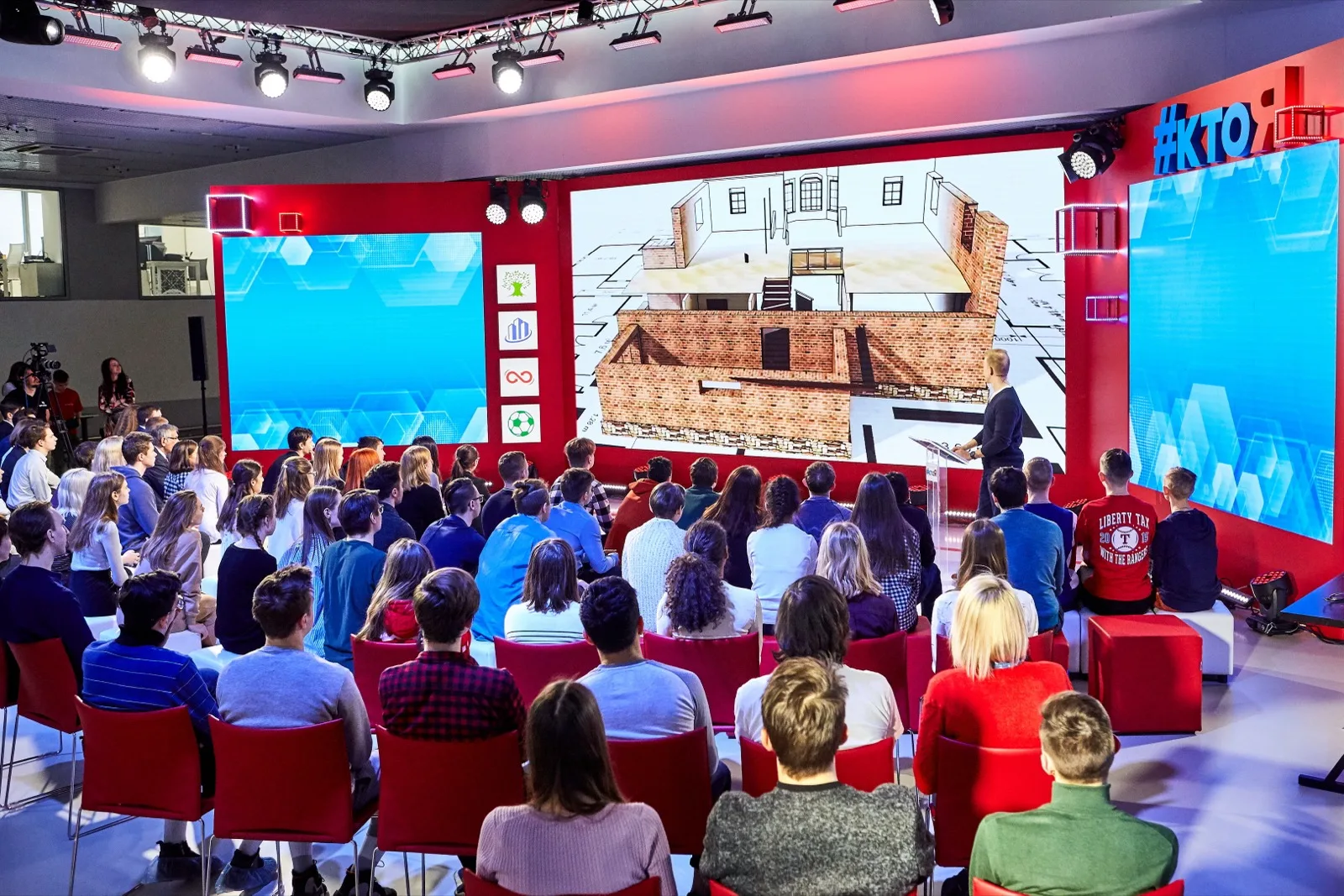 Wide shot of the studio: three screens, the audience, and the host at the podium